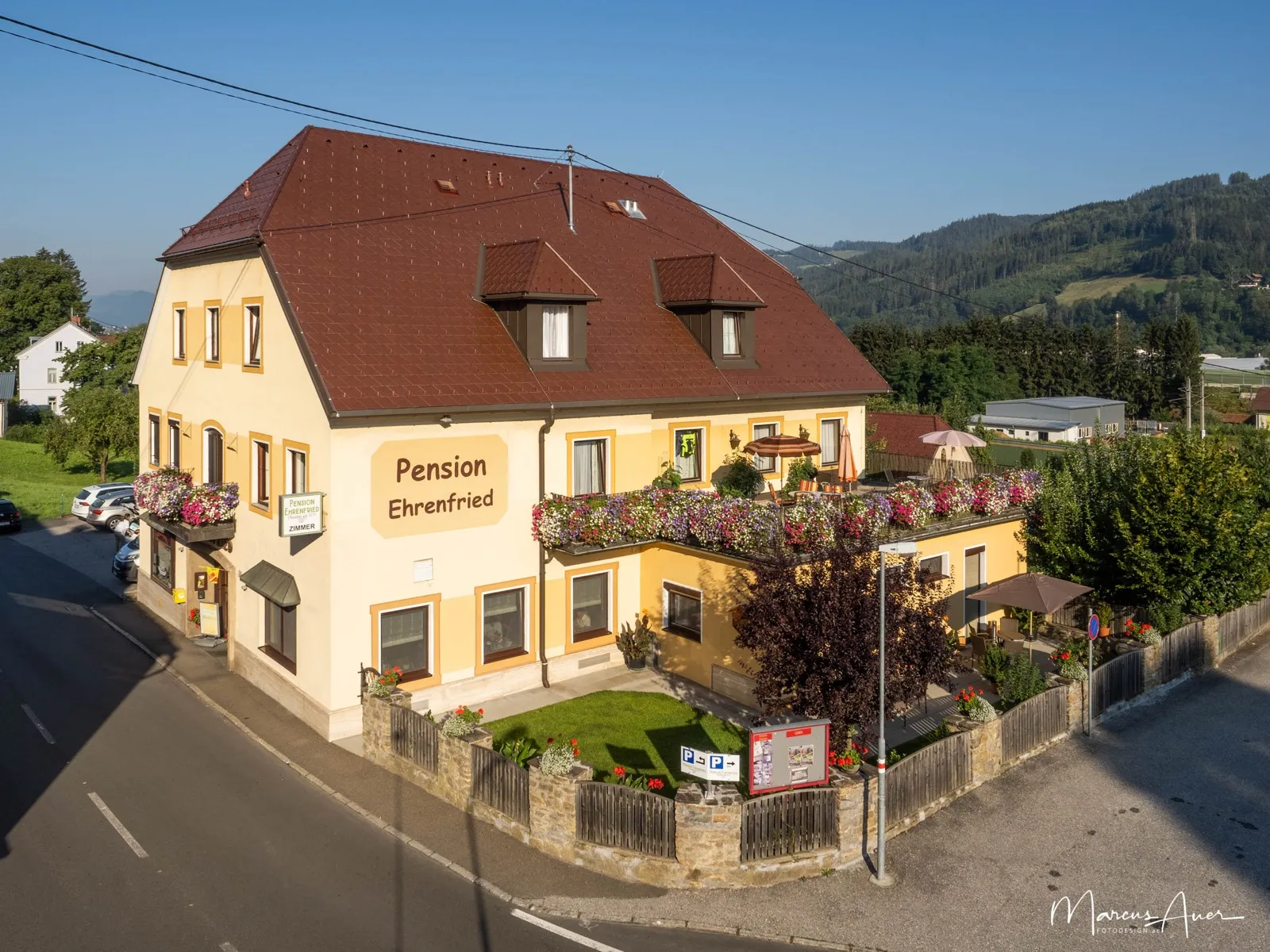 Außenansicht der Pension Ehrenfried mit gelber Fassade und Blumenschmuck, im Hintergrund die grünen Hügel der Hochsteiermark